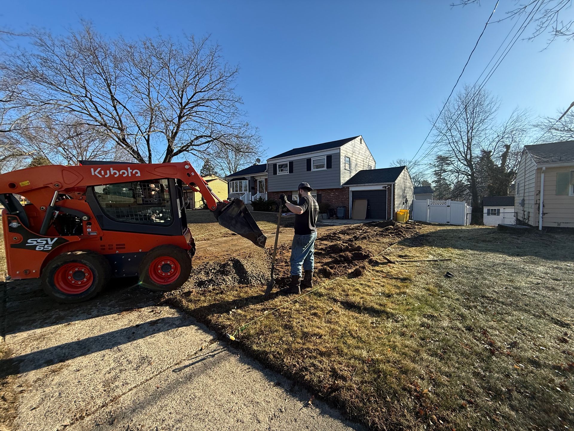 Worker near orange utility lift in yard beside house, with bare trees and fallen leaves.
