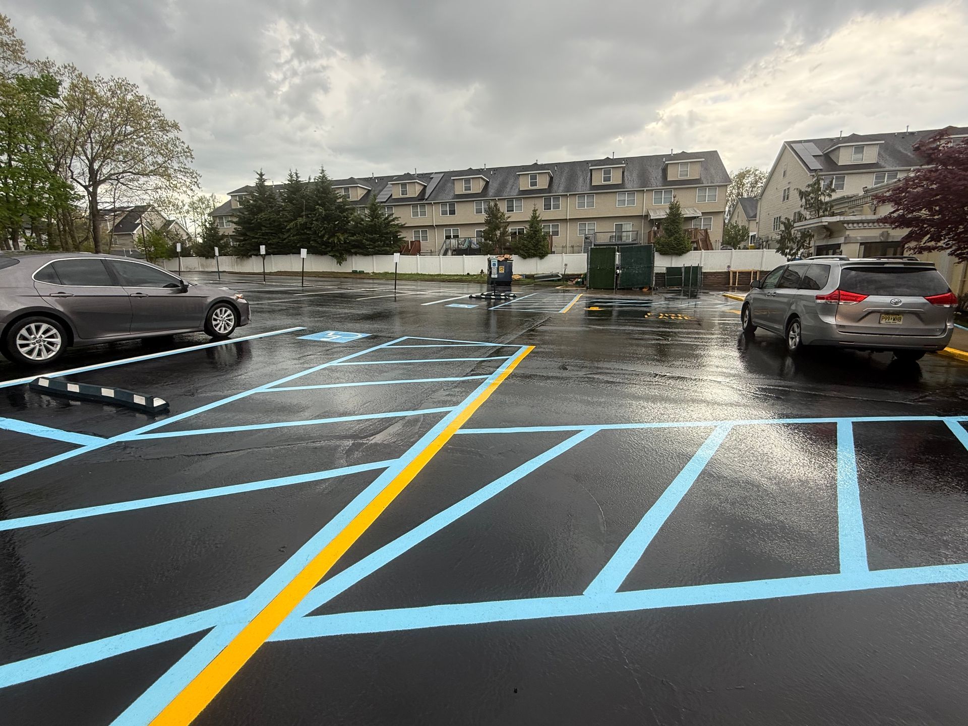Rain-soaked parking lot with blue and yellow lines, parked cars, and apartment buildings under gray skies