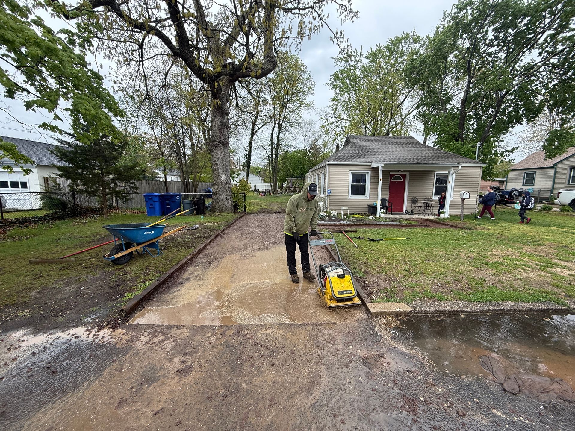 Person operating a yellow plate compactor on a muddy driveway in front of a small house.