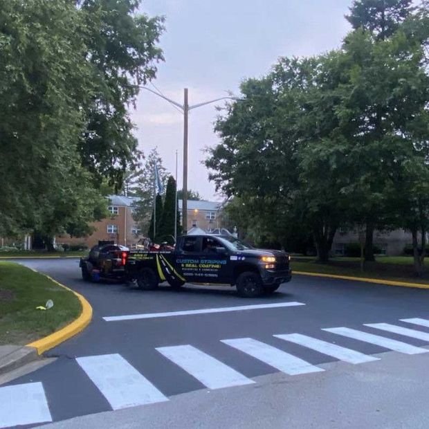 A tow truck towing a car across a crosswalk with buildings and trees in the background.
