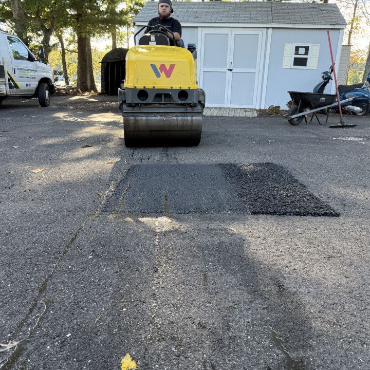 Person operating a yellow asphalt roller compacting fresh asphalt on a driveway.
