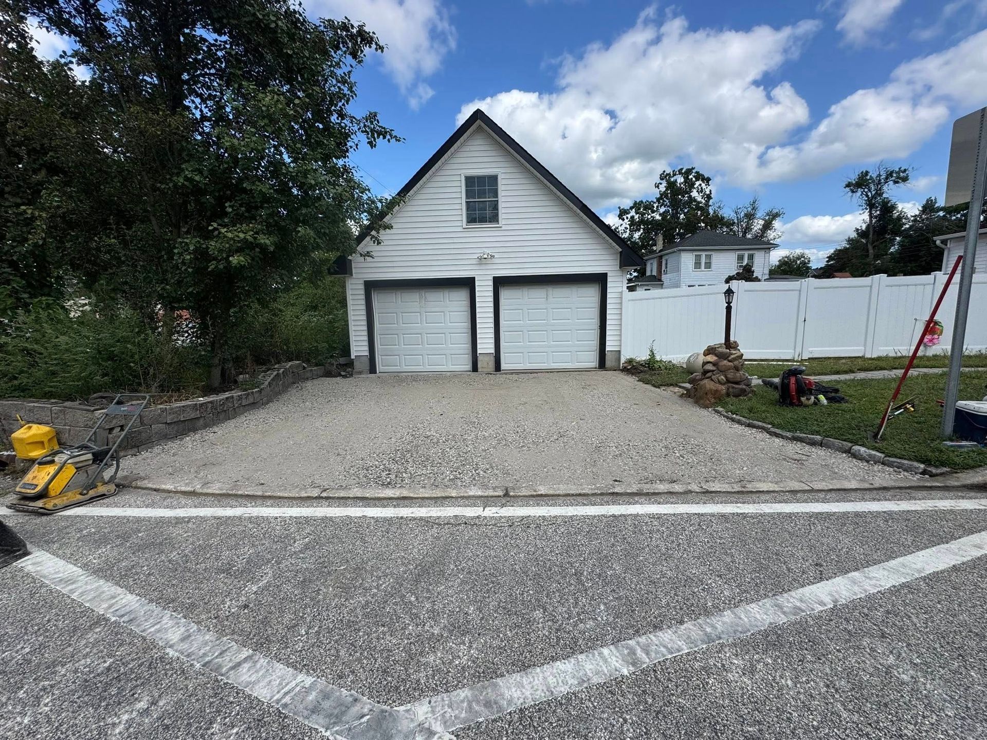 Gravel driveway leading to a small white garage with two doors and a white fence behind it.