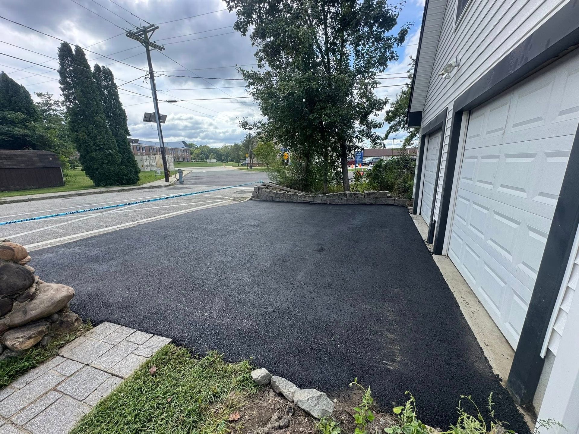 Fresh black asphalt driveway beside a white garage, with a street, trees, and cloudy sky beyond.