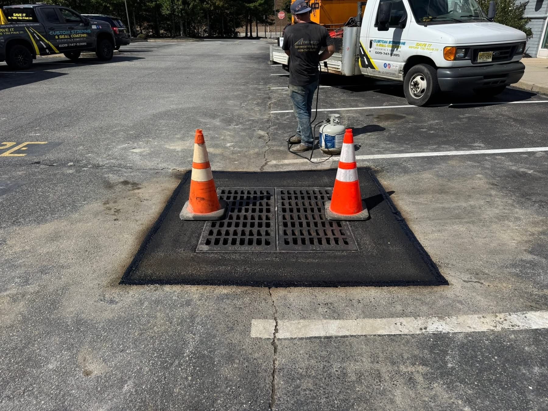 Traffic cones around a storm drain grate in a parking lot, with a worker and utility truck nearby.