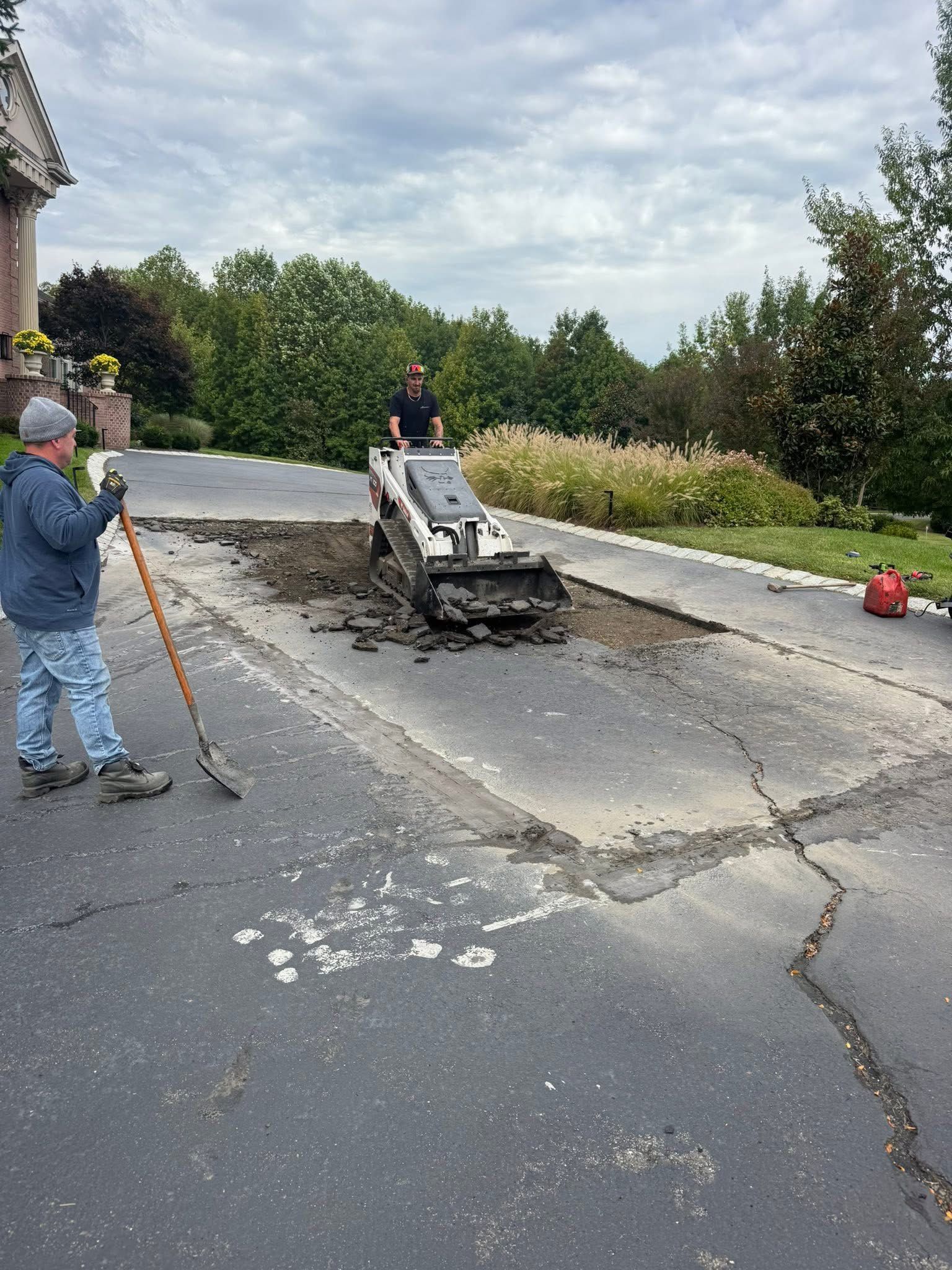 Workers repair a cracked driveway with a small paving machine and tools outdoors.