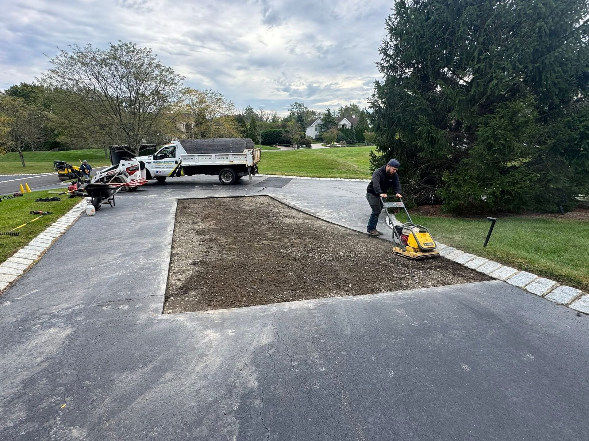 Driveway repair scene with workers, a dump trailer, and fresh asphalt patching around a tree.