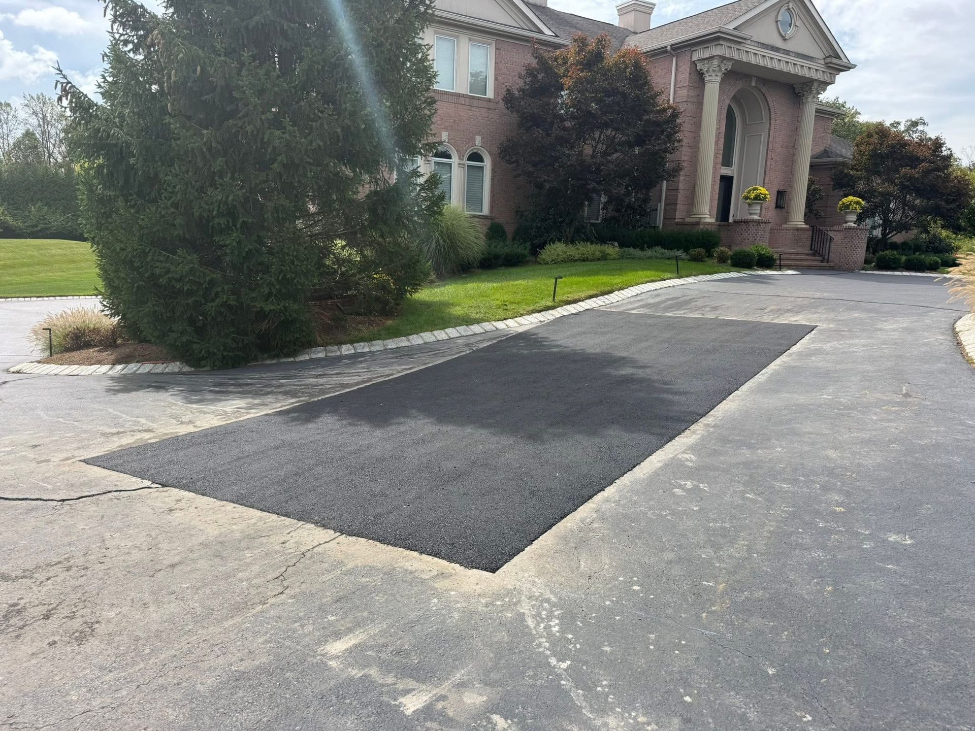 Fresh asphalt patch on a driveway in front of a house, with trees and shrubs nearby