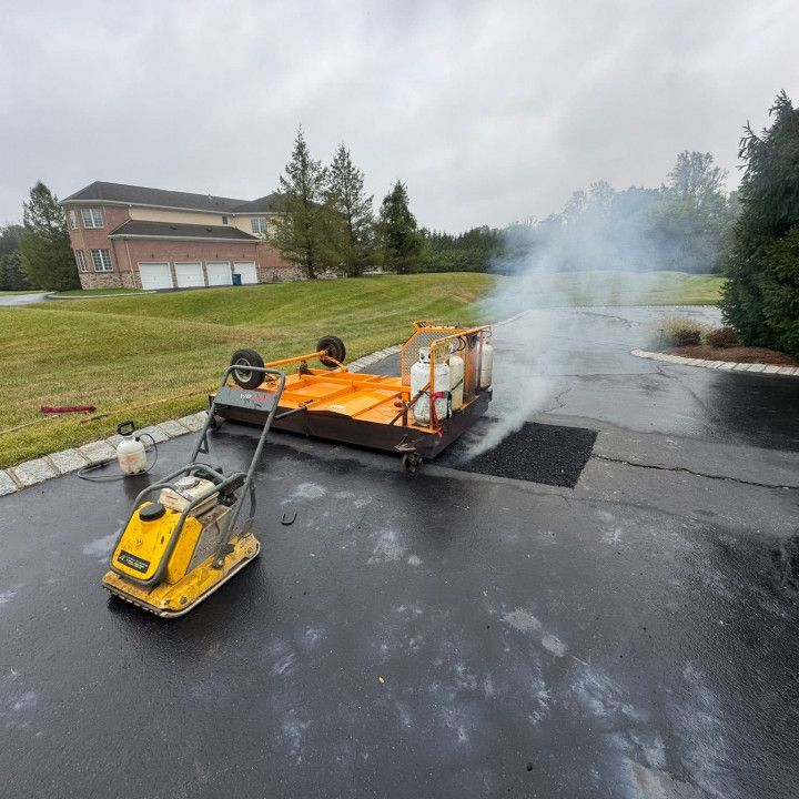 Asphalt repair on a driveway: Steam rising from a small patch. Equipment includes a vibratory plate compactor and a propane heater.