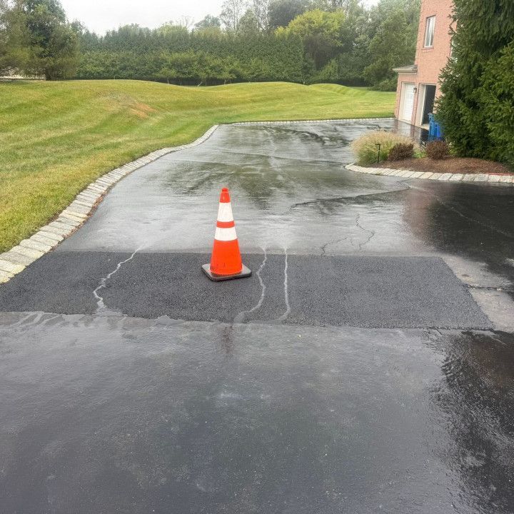 Orange traffic cone on wet, cracked asphalt driveway. Driveway leads to house and lawn.