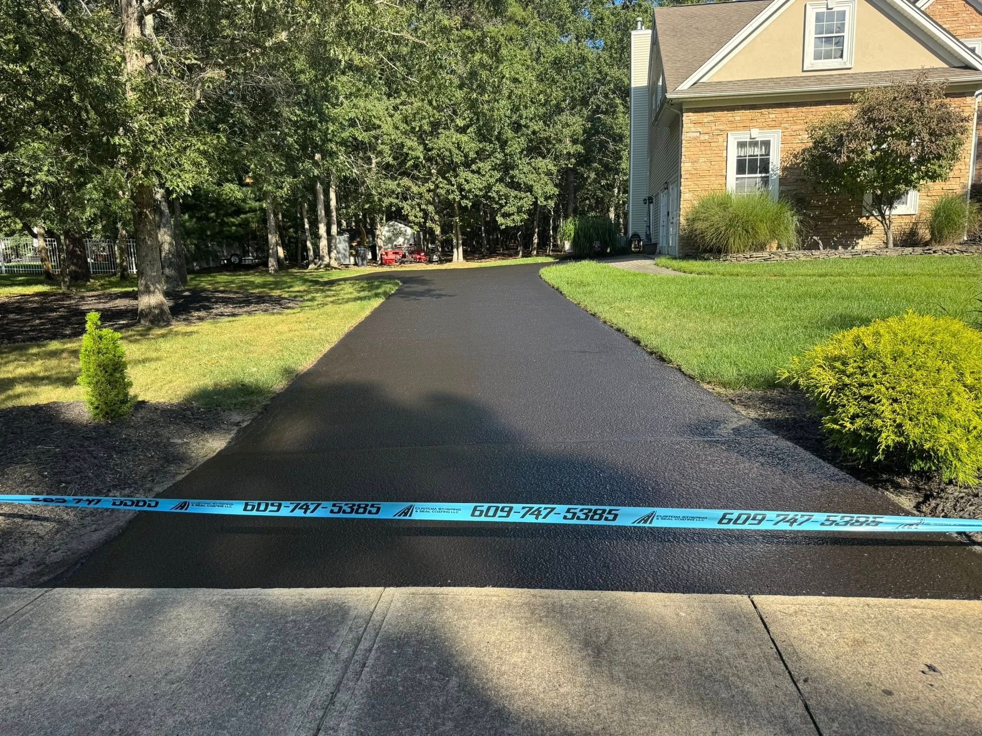 Freshly paved black asphalt driveway with blue caution tape, flanked by grass and trees leading to a house.