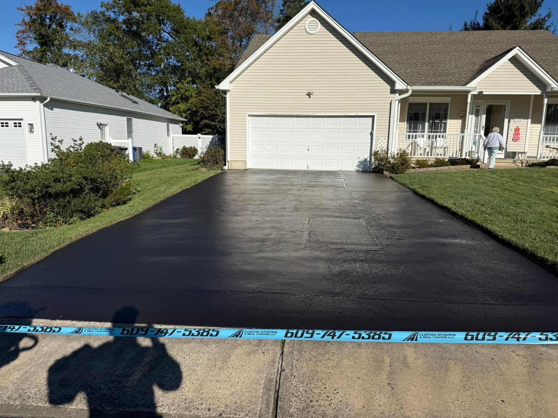 Freshly sealed black driveway in front of a tan house with a garage; blue tape in the foreground.