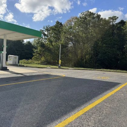 Gas station with green canopy, asphalt, and yellow lines. Trees and a cloudy blue sky in the background.