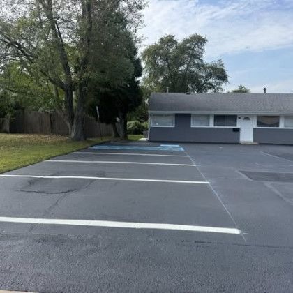 Parking lot with painted lines in front of a gray building and green grass, with trees in the background.