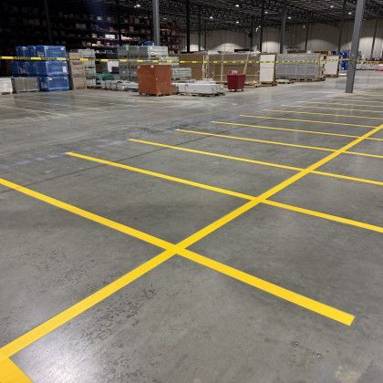 Yellow lines painted on a concrete warehouse floor, with boxes and shelving in the background.