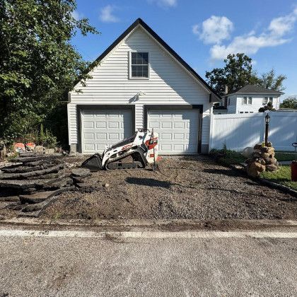 Driveway renovation in progress: white garage with bobcat, removed asphalt, gravel, blue sky.