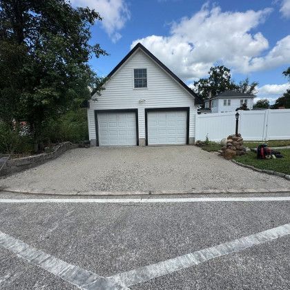 White two-car garage with gravel driveway. Asphalt road in the foreground, cloudy sky.