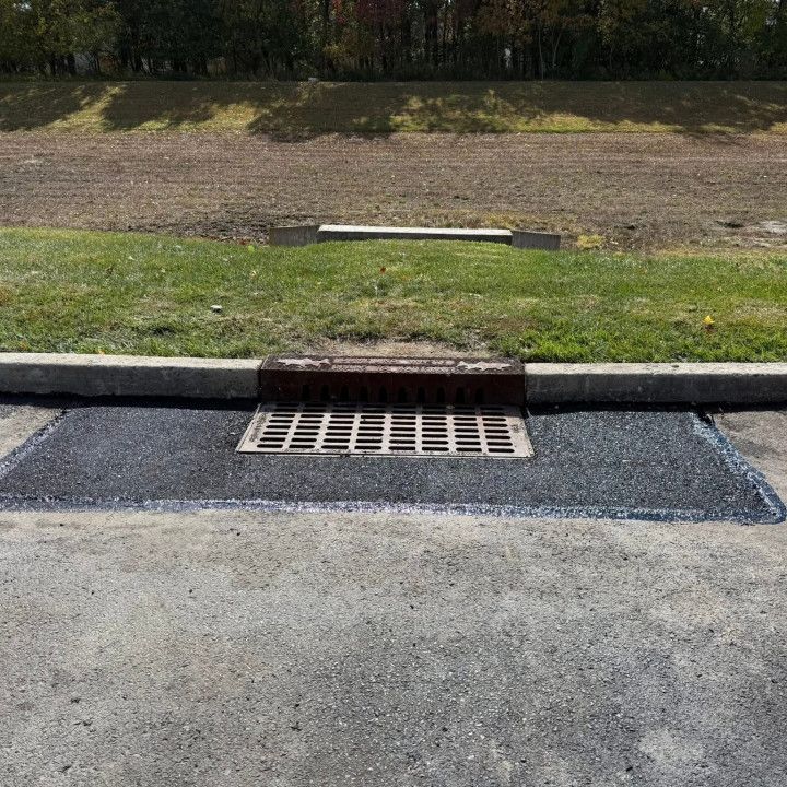 A metal drain with asphalt around it in a parking lot, near a grassy area and curb.
