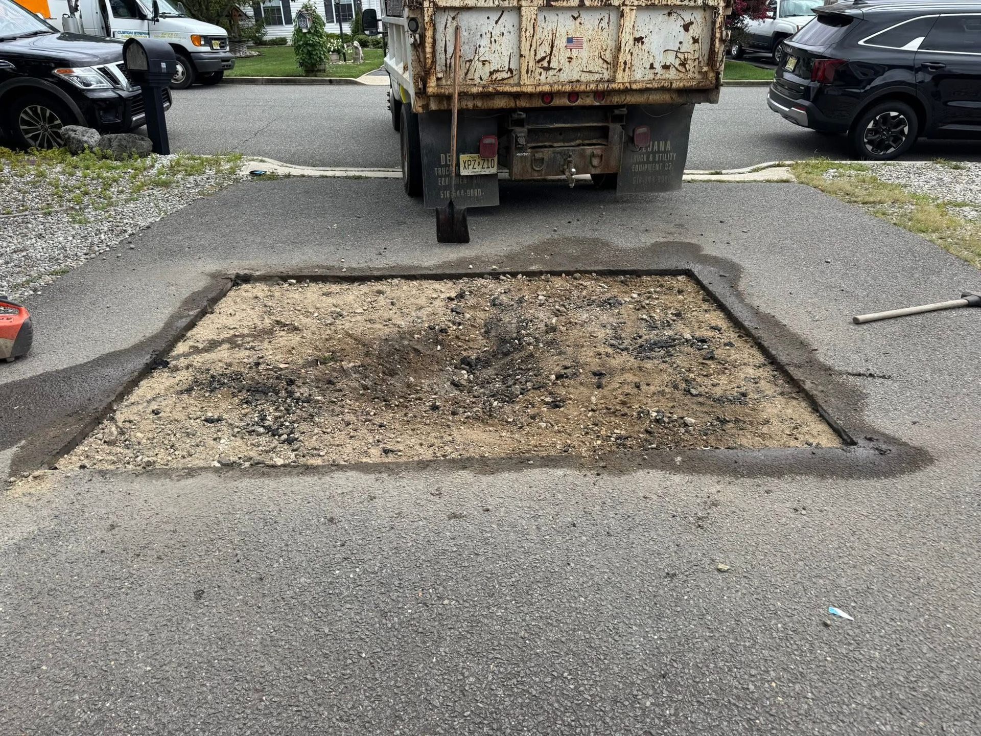 Pothole-style road repair patch in a parking lot behind a rusty truck, with parked cars nearby