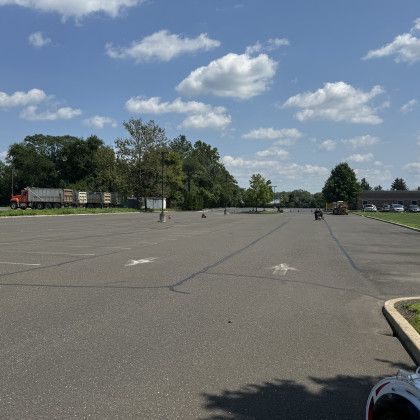 Empty asphalt parking lot under a blue sky with fluffy clouds; trees and buildings in the background.