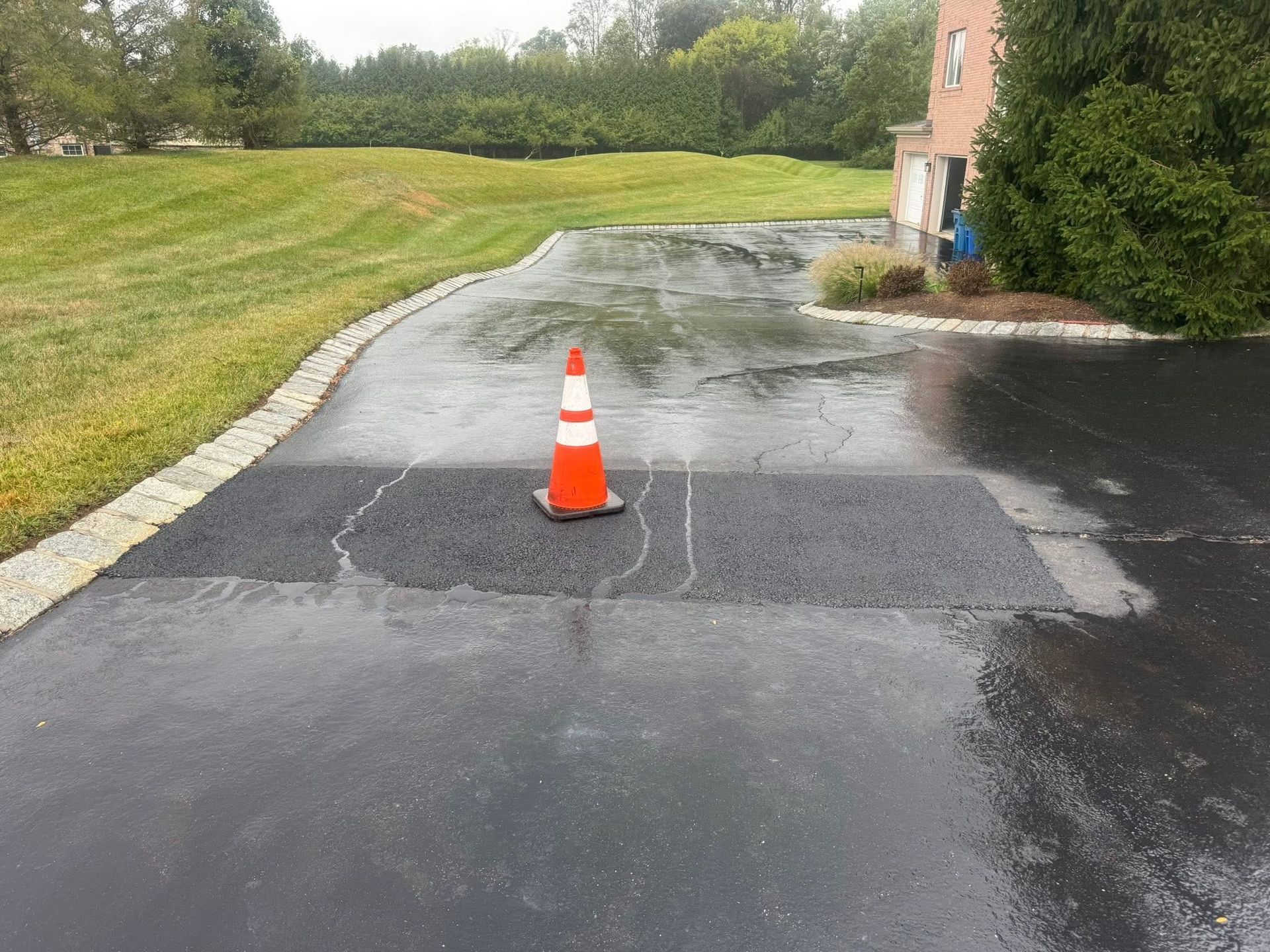 Orange traffic cone on a wet driveway beside a brick building and grassy lawn.