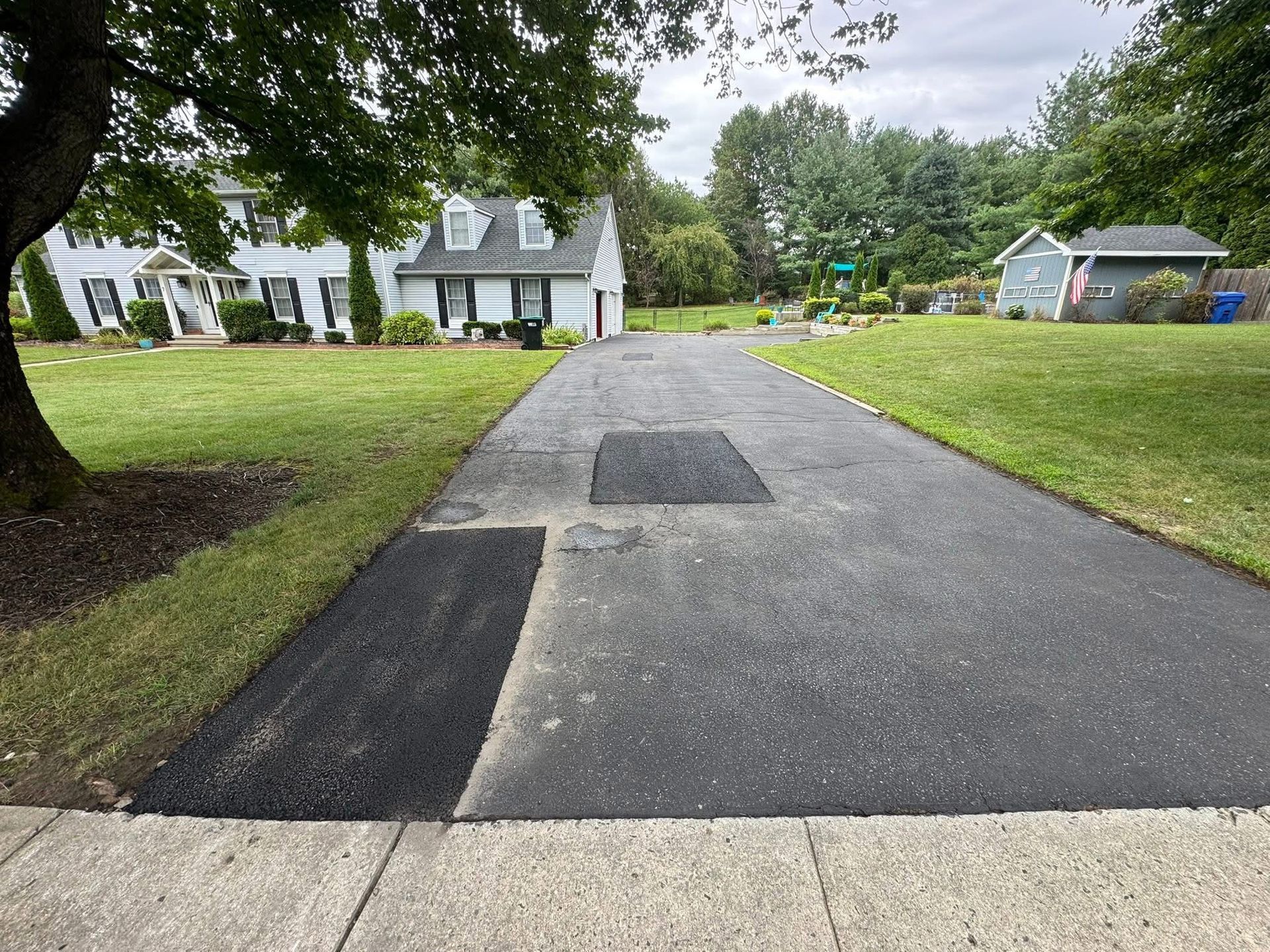 Freshly paved driveway with dark asphalt patches leading to a suburban house on a grassy lot