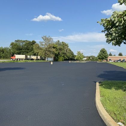 Newly paved asphalt parking lot on a sunny day; several trees and a truck visible in the background.