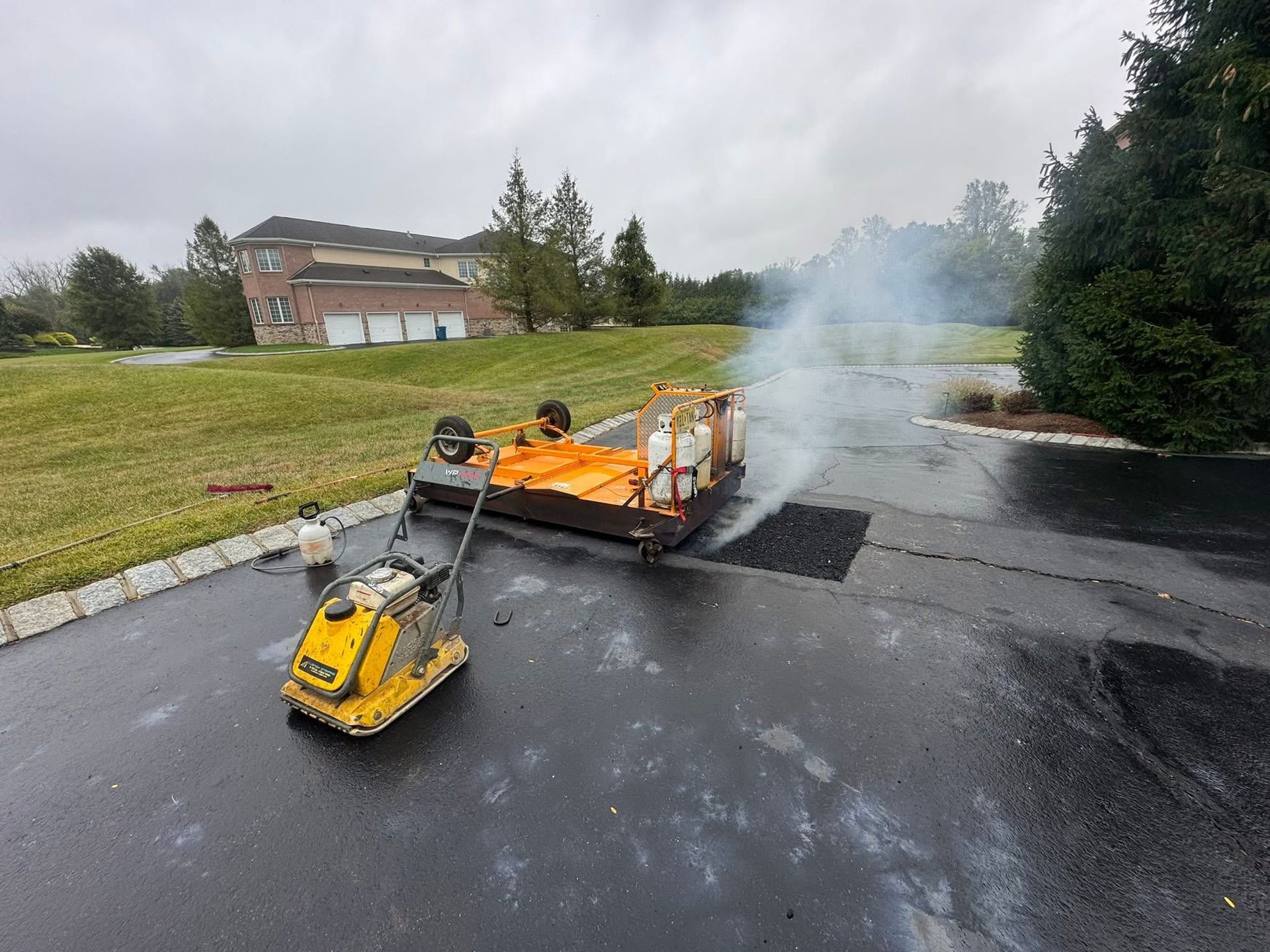 Yellow road machine towing an orange trailer on a wet road, with smoke rising nearby.