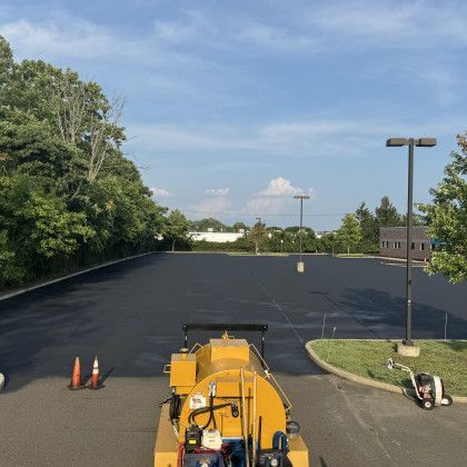 Newly paved black parking lot; asphalt machine in foreground, trees and buildings in background.