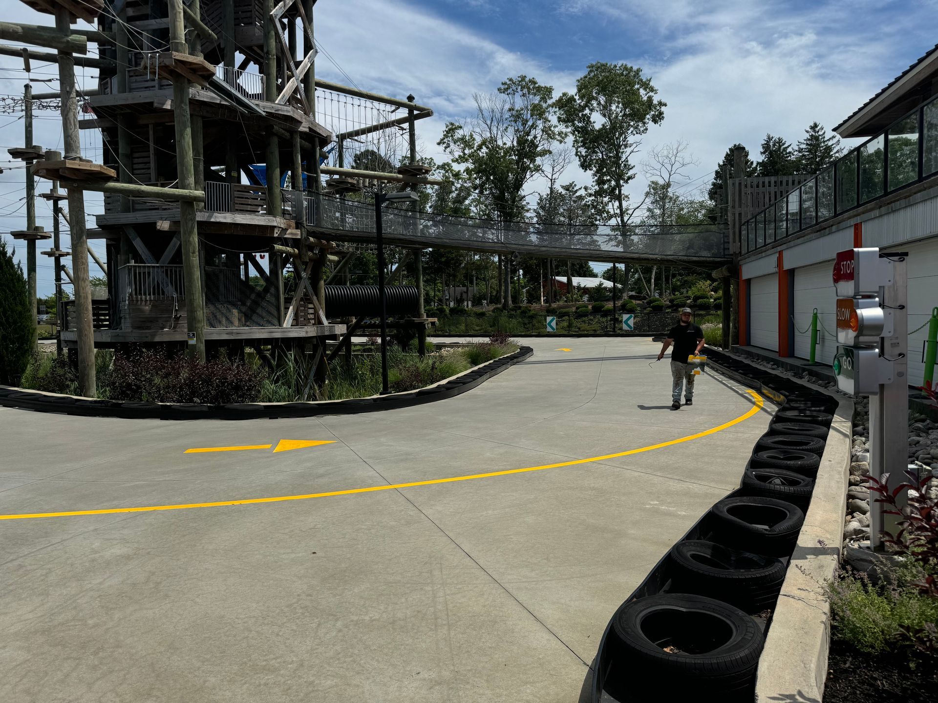 Man walking on a go-kart track with tire barriers, an adventure course in background, and a sunny sky.