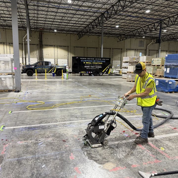 A person using a floor grinding machine in a warehouse. They wear a safety vest and a hat.