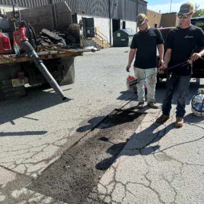 Two workers repairing asphalt with a torch and tools near a truck.
