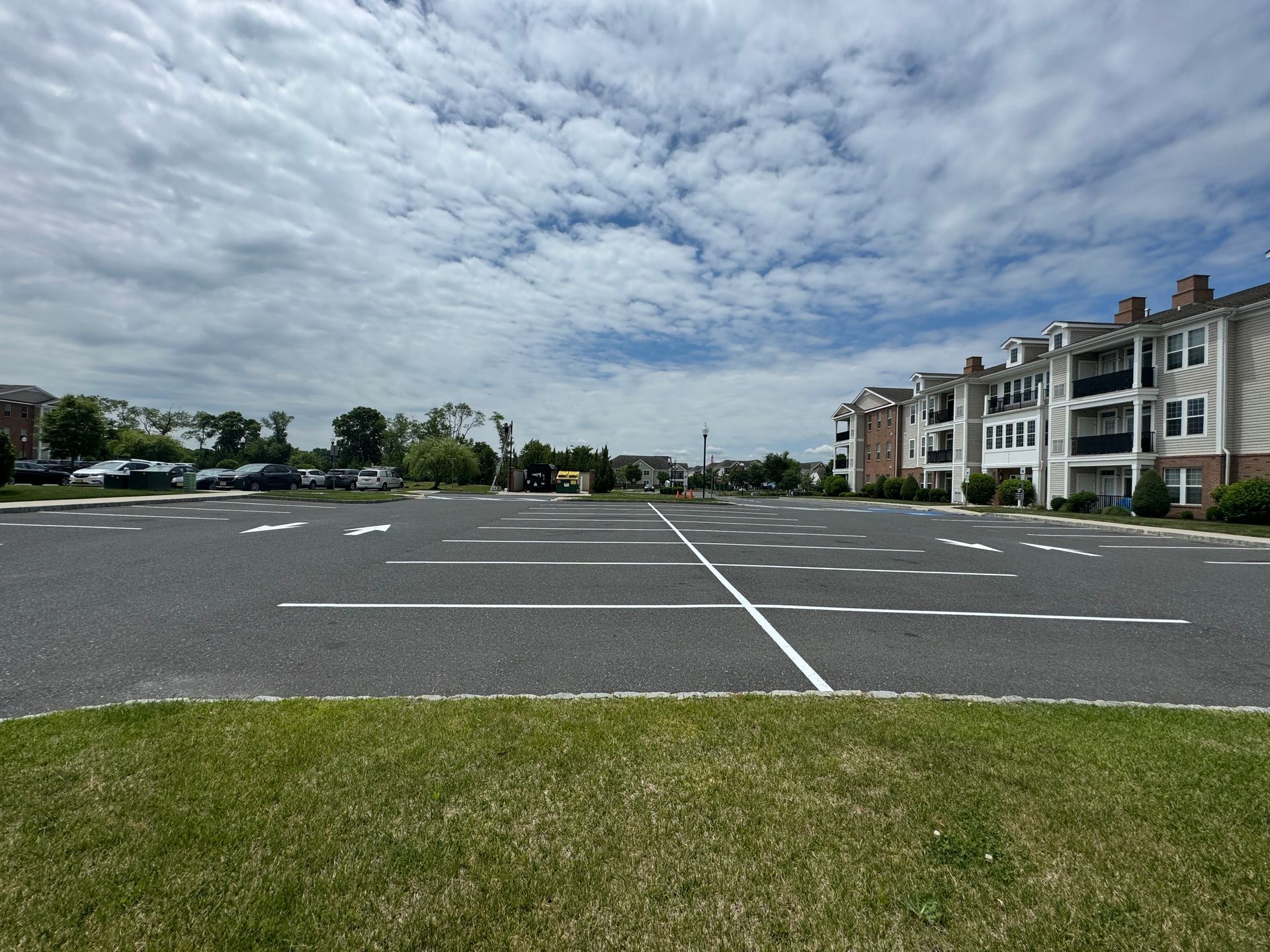 A large, empty parking lot with white lines, bordered by grass and apartment buildings under a cloudy sky.
