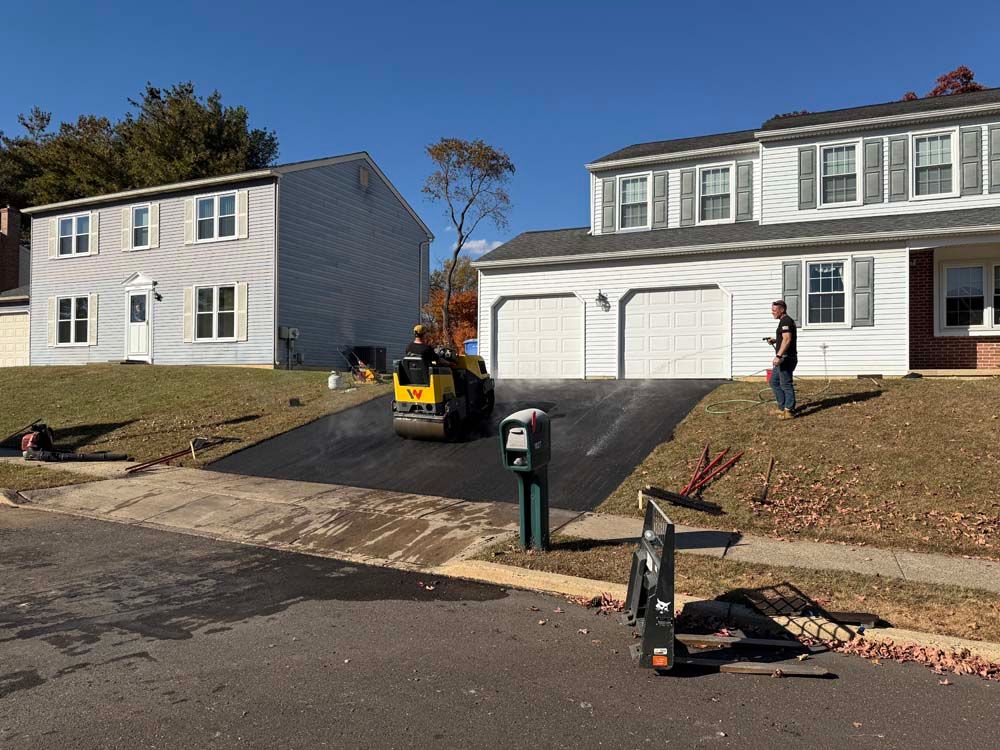 Workers and a yellow skid steer repair a driveway between two suburban houses on a sunny day