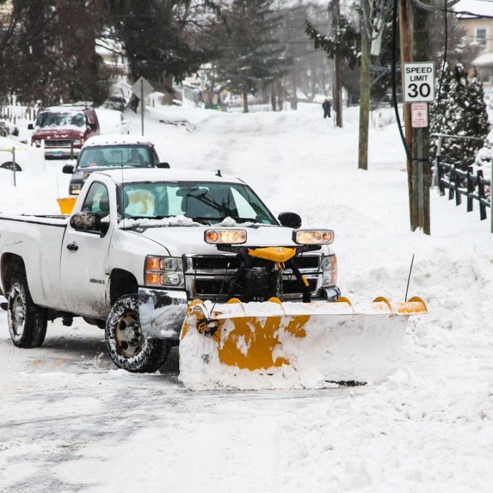 White pickup truck with a snowplow clearing a snow-covered street; other vehicles and a speed limit sign are visible.
