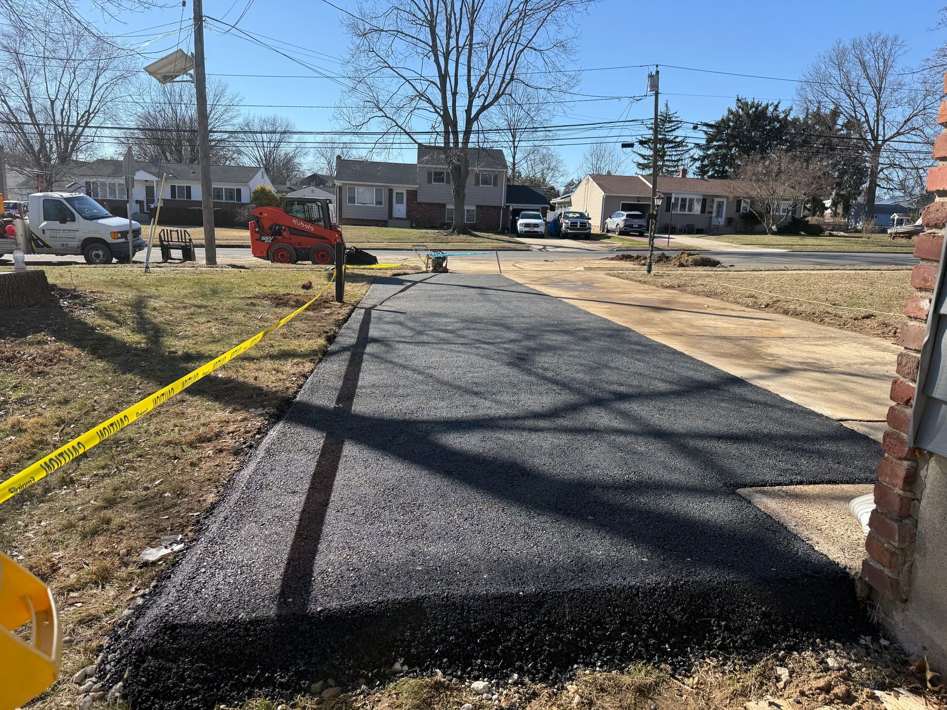 New blacktop driveway under construction beside a brick house, with a yellow caution tape and workers in the distance