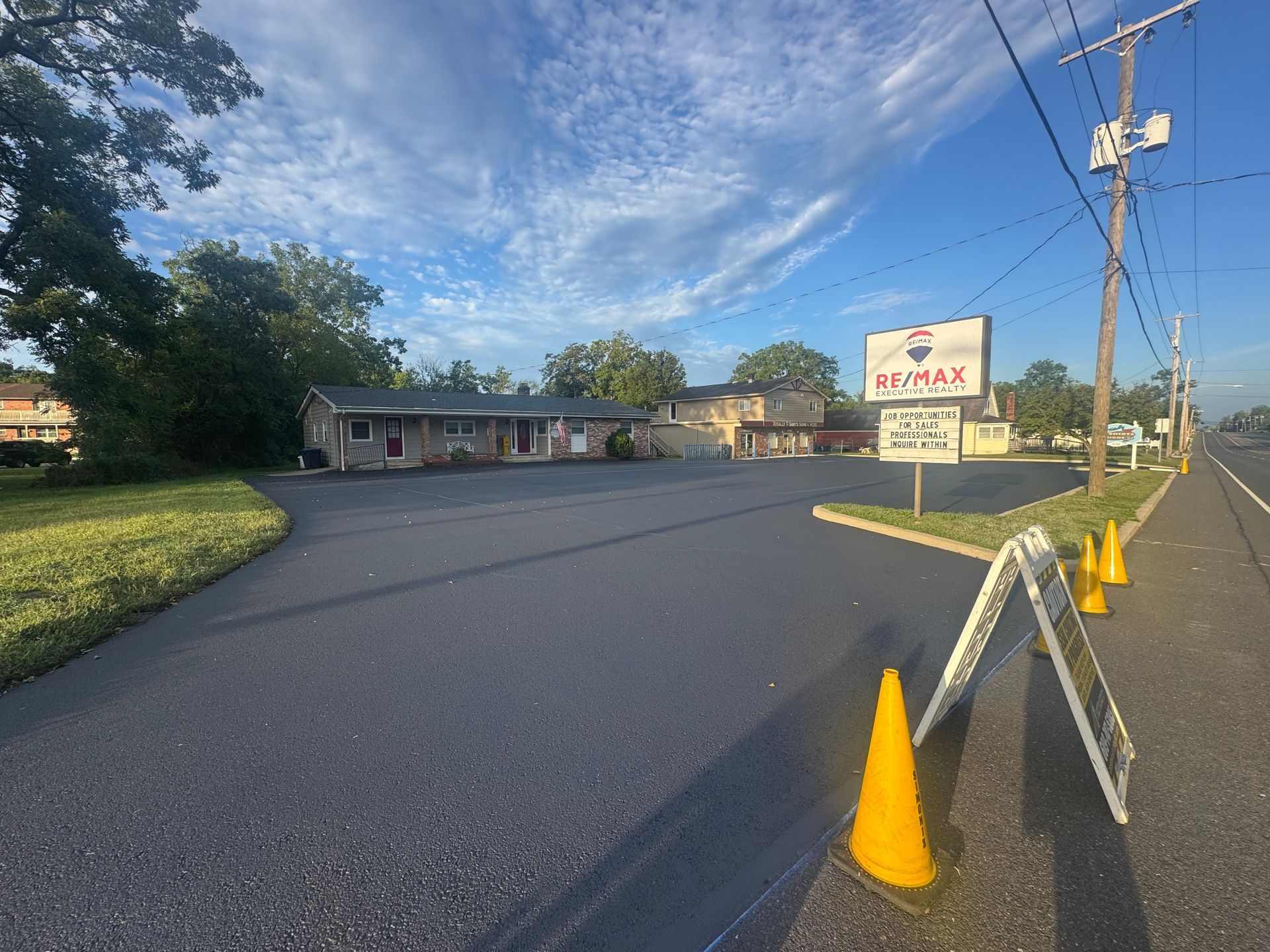 Paved parking lot with business sign, two A-frame signs, and traffic cones along a road.