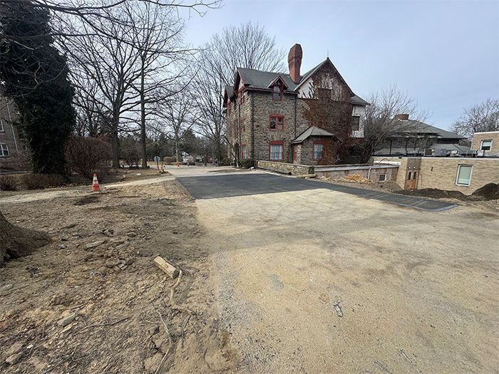 A gravel road with a freshly paved section leading toward a stone house with bare trees.