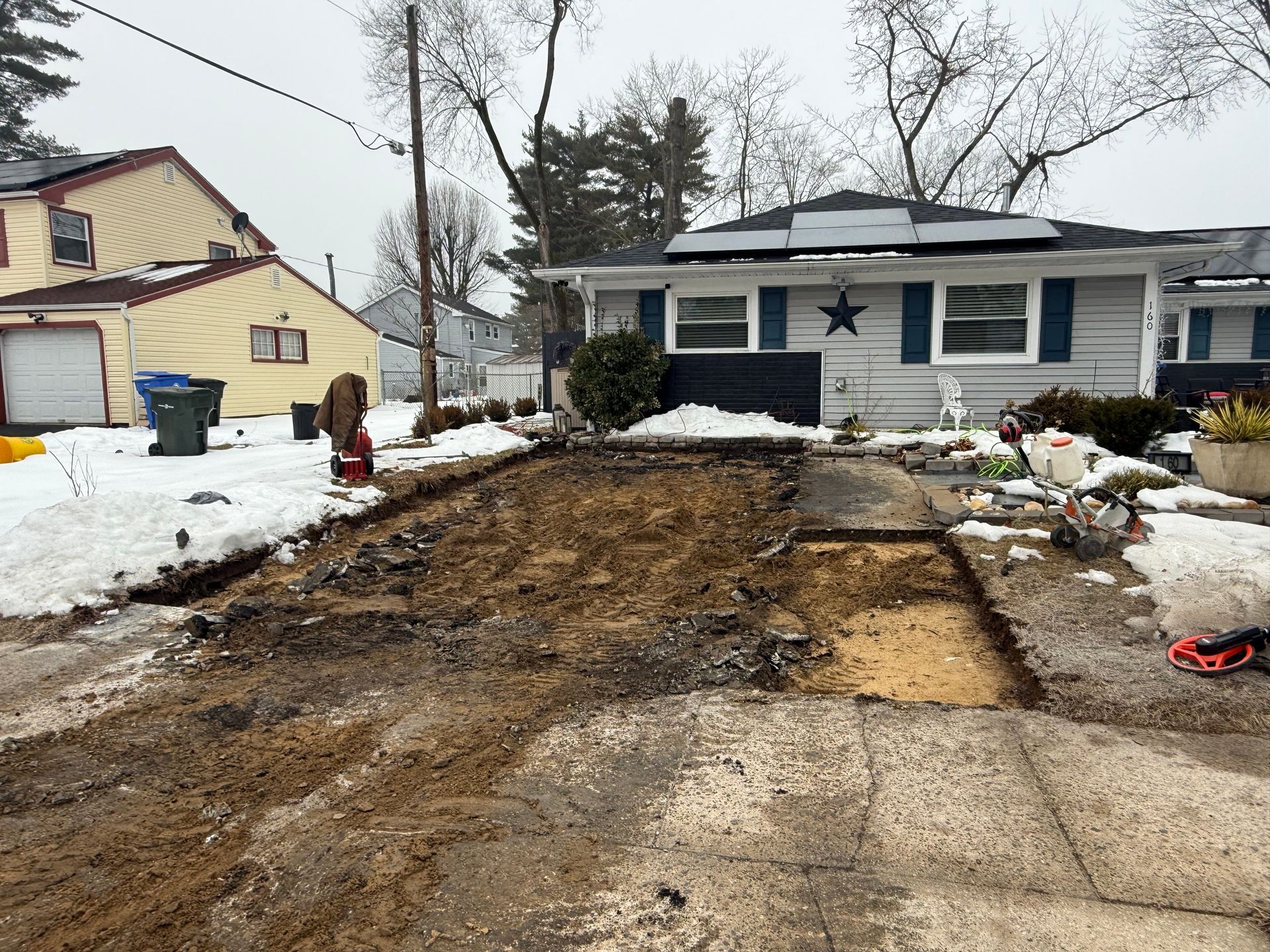 Mud and torn pavement in a snowy residential driveway beside a blue house under repair.