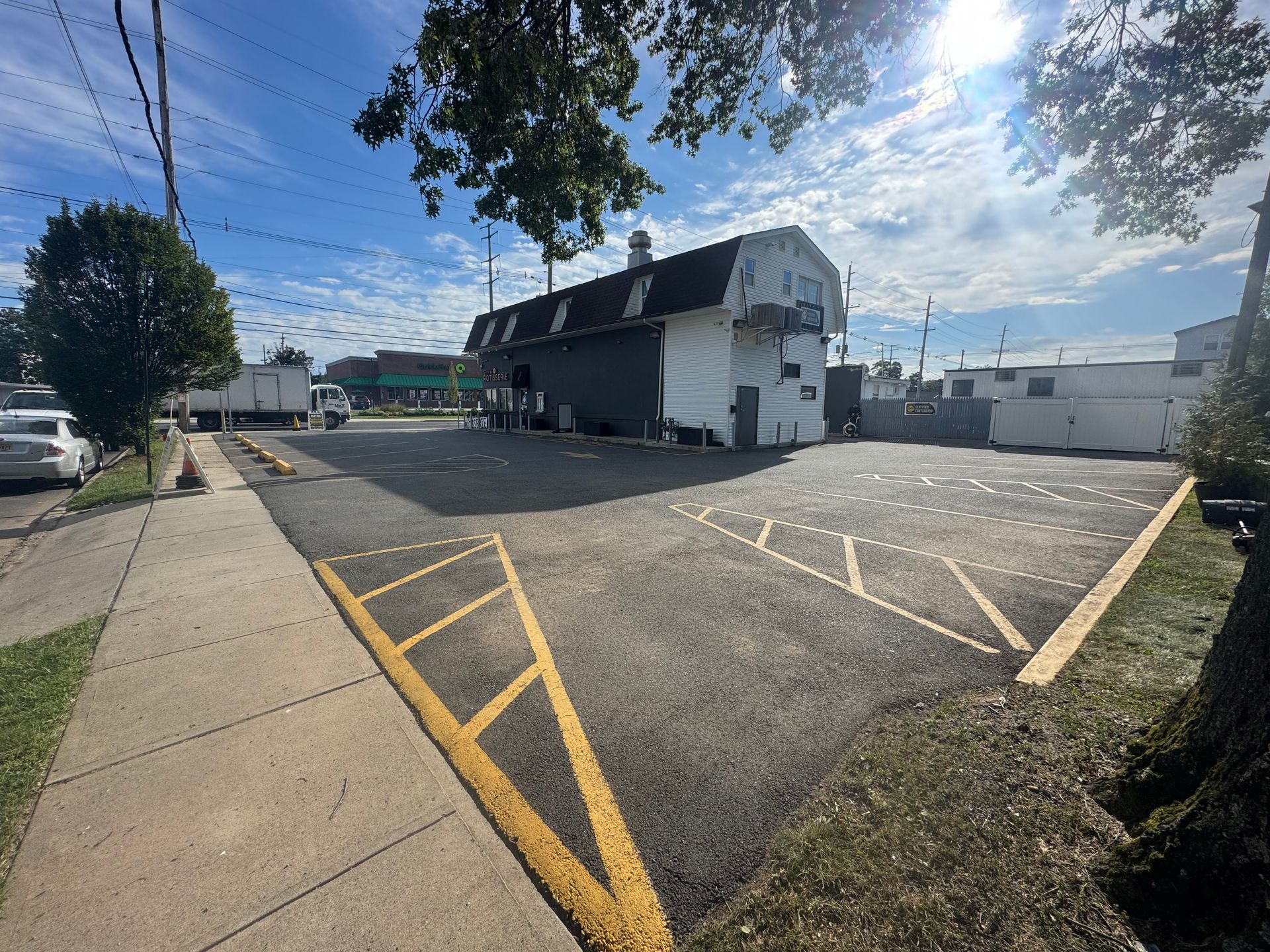 Paved parking lot with yellow markings, building with basketball hoop, sidewalk, and street. Blue sky and trees.