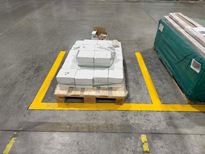 Pallet of white packages within a yellow painted storage area on a gray warehouse floor. Another wrapped item is to the right.