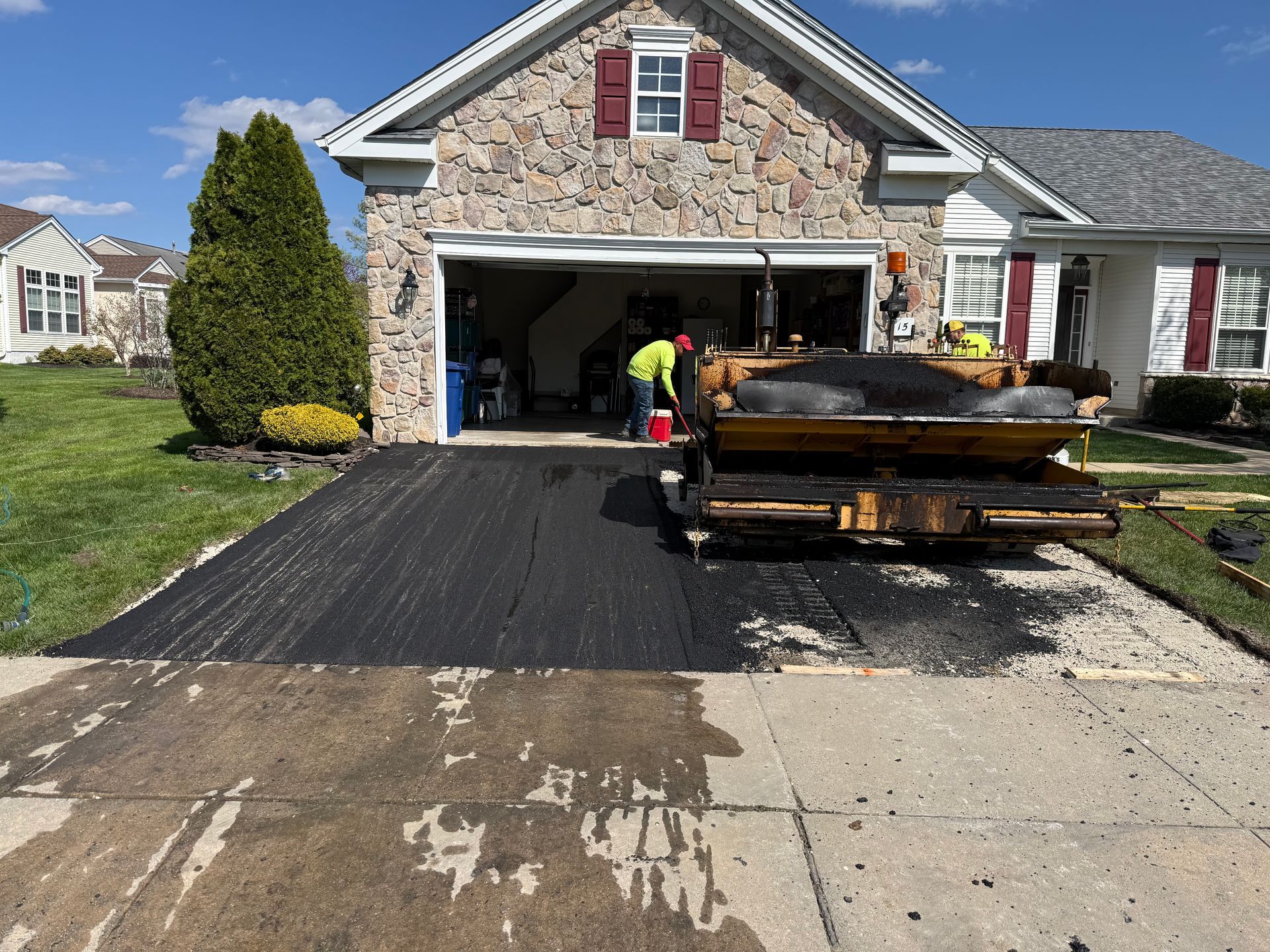 Driveway resurfacing with fresh black asphalt and a truck outside a stone house garage.