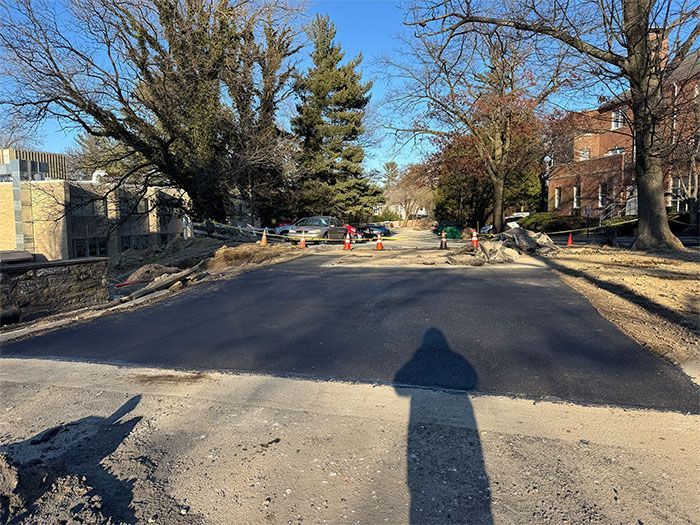 Newly paved asphalt road; construction site with cones; houses and trees in background.