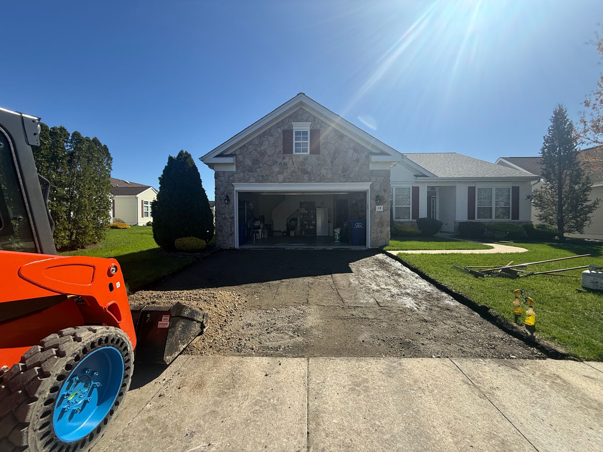 House with open garage and unfinished driveway; orange tractor in foreground under bright sun.