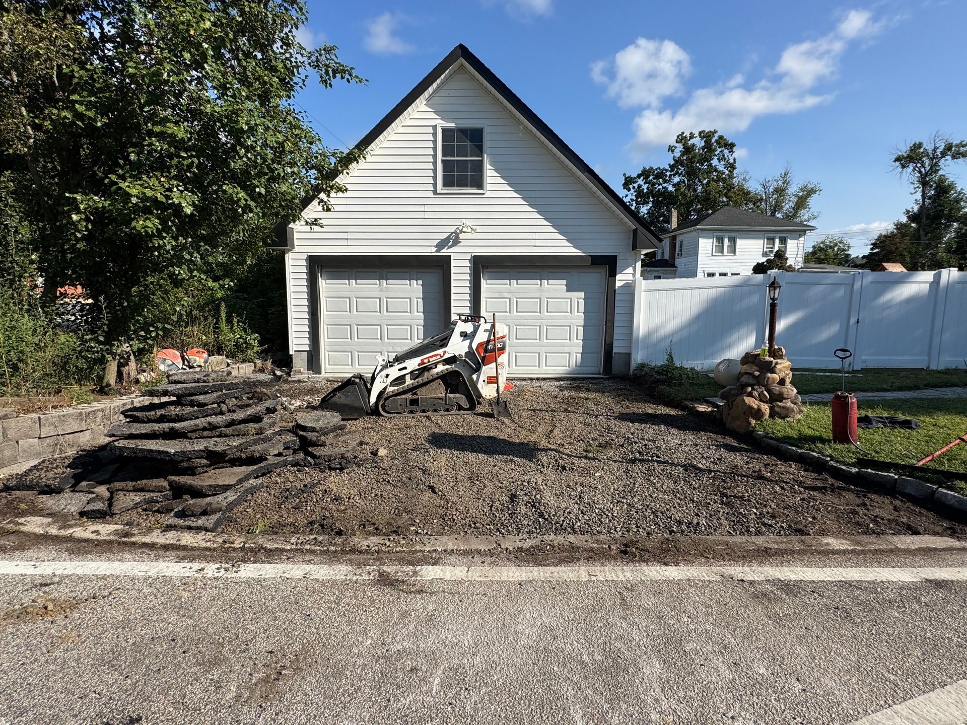 Driveway under construction with a small bulldozer, in front of a white garage under a blue sky.