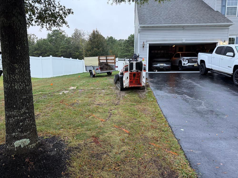 Lawn equipment parked on wet driveway beside open garage and parked trucks, with white fence and tree nearby