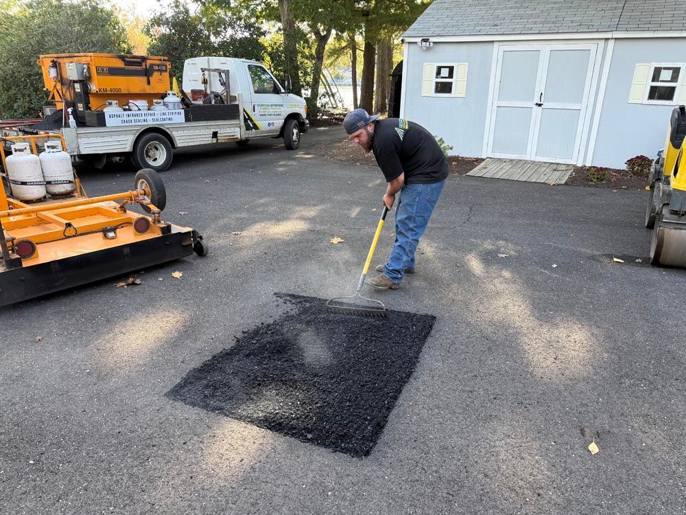 Worker patching asphalt in a driveway beside a white utility truck and orange equipment.