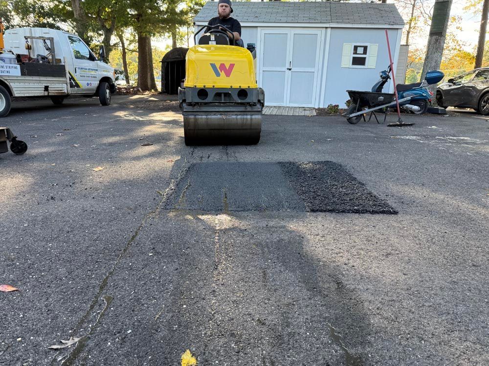 Road roller paving a patched asphalt driveway beside a white shed with vehicles parked nearby
