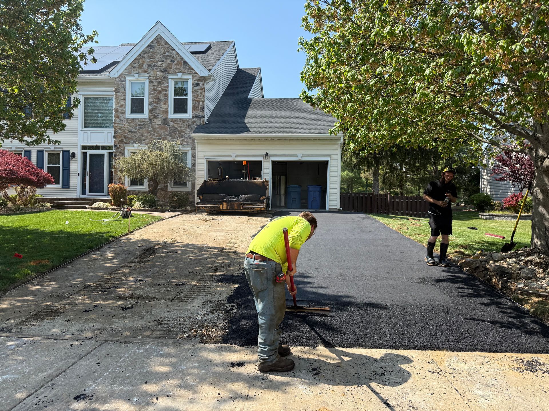 Workers paving a driveway at a house, one in a neon vest spreading asphalt near a tree-lined yard.