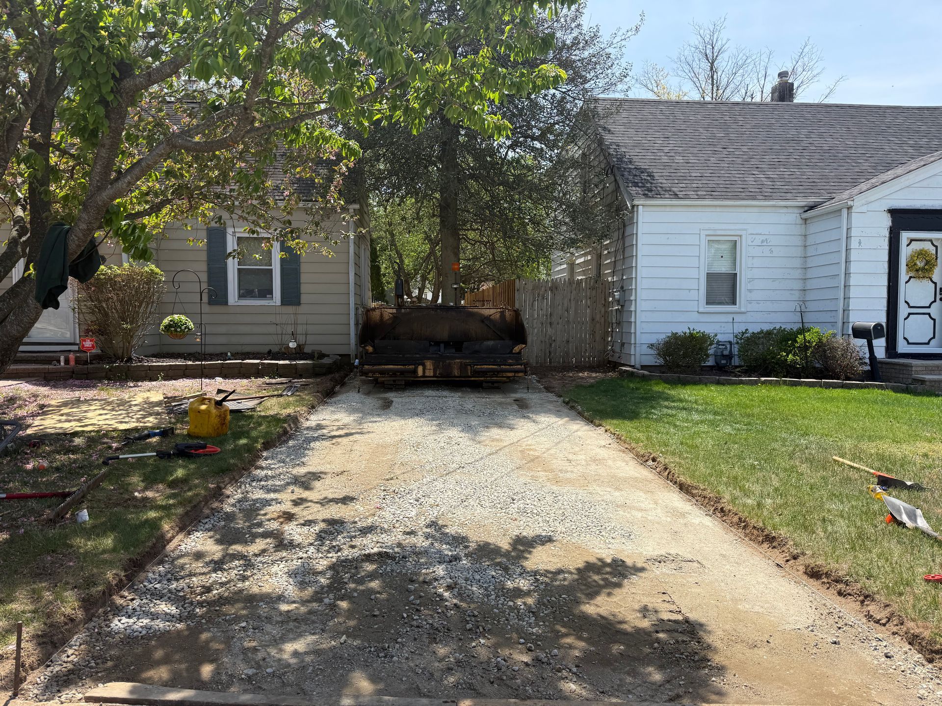 Driveway between a tan house and white garage, with a tree casting shade and a green lawn.