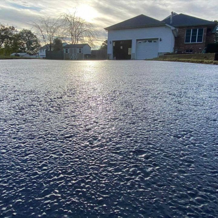Black driveway reflects sunlight, leading to a house with a garage and another building in the distance.